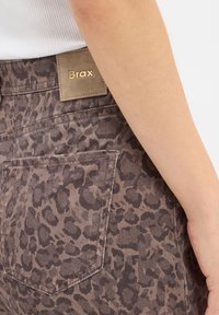 Pantalons à imprimé léopard marron en tissu texturé, avec une grande poche arrière et une étiquette beige portant "Brax" en lettres dorées.