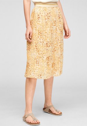 Žlutá plisovaná sukně s květinovým vzorem po kolena, nošená s béžovými žabkami na stojící osobě na neutrálním pozadí.