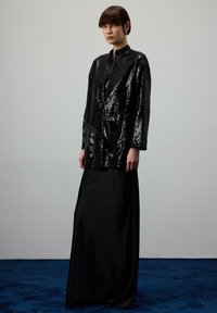 Čierna sako s flitrami a opaskom v páse, doplnená elegantnou čiernou maxi sukňou. Oblečenie dominuje hladkou textúrou a trblietavými detailmi.