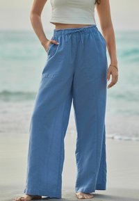 Pantaloni largi de culoare albastru deschis, dintr-un material moale, cu un talie elastică și buzunare laterale, purtați cu un top scurt, stând pe nisip.