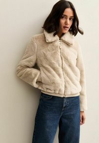 Chaqueta beige de pelo sintético con cuello, cremallera frontal y puños acanalados. Textura suave, ajuste holgado y bolsillos laterales. Se lleva con vaqueros oscuros.