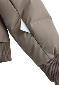 Barna pufi kabát ujja bordázott mandzsettával, oldalzsebbel, amelyben egy fehér kártya látható. Simább, matt textúra.