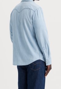 Jasnoniebieska koszula dżinsowa z długimi rękawami, mankietami na guziki i zaokrąglonym dołem. Gładka faktura z klasycznym kołnierzem i detalem tylnej części yoke.