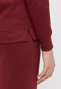 Burgundowa bluza z ściągaczami przy mankietach i dolnym brzegiem, z bocznym rozcięciem dla wygody ruchu. Miękka, fakturowana tkanina.