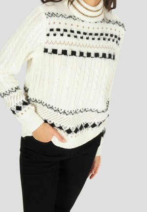 Vrouw draagt een witte kabelgebreide trui met zwarte geometrische patronen en een zwarte broek, hand in de zak, tegen een effen achtergrond.