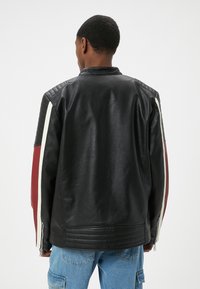 Giacca di pelle nera con spalline testurizzate, accenti a righe rosse e bianche sulle maniche e polsini con zip. Finitura liscia e brillante.