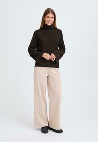 Brauner gerippter Strickpullover mit hohem Kragen und ausladenden Ärmeln, kombiniert mit beigen weiten Hosen und schwarzem Schuhwerk. Einfaches, modernes Design.