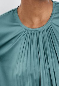 Top plissettato color teal realizzato in un tessuto morbido; presenta un colletto rotondo e una leggera caduta, con pieghe verticali che si estendono dal neckline.