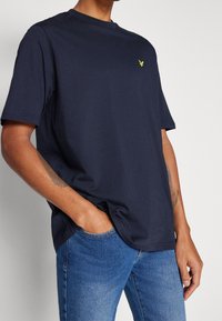 Granatowa bawełniana koszulka z krótkim rękawem, okrągłym dekoltem i małym żółtym logo orła na klatce piersiowej. W połączeniu z niebieskimi dżinsami.