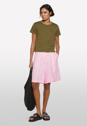 T-shirt cropped verde oliva, pantaloni corti in lino rosa chiaro con pieghe e sandali neri bassi. Modella tiene una borsa tote in rete nera.