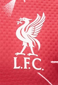 Λευκό έμβλημα Liver bird που κρατάει ένα κλαδί πάνω από τα γράμματα «L.F.C.» σε κόκκινο υφασμάτινο φόντο με υφή.