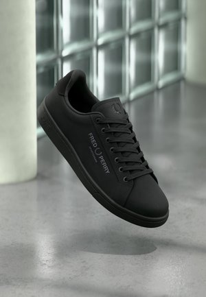 Schwarzer Fred Perry Sportswear-Sneaker mit Schnürsenkeln, Logo an der Seite und auf der Zunge, präsentiert vor einer modernen Glasblockwand und einem Betonboden.