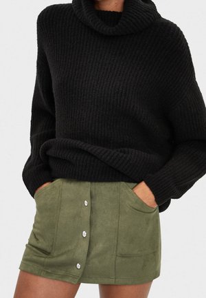 Sort chunky strikket sweater med rullekrave kombineret med olivengrøn kort ruskindnederdel med knapper foran og lommer, hænderne placeret i lommerne.