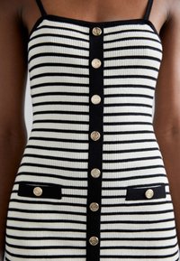 Robe sans manches à rayures horizontales noires et blanches avec une bande centrale noire et des boutons dorés sur le devant et sur les rabats des poches.