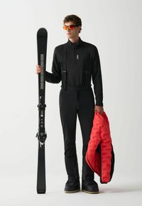 Uomo in tuta da sci nera e occhiali da sole arancioni, che tiene uno sci in una mano e una giacca rossa nell'altra su uno sfondo neutro.