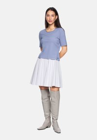 Helles blaues Kurzarmoberteil kombiniert mit einem weißen, gefalteten Rock mit silbernen Streifen, ergänzt durch knielange graue Lederstiefel.