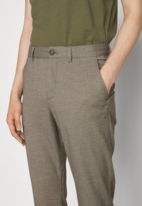 Brązowe spodnie w kratę z dopasowanym stanem, bocznymi kieszeniami i zapięciem na guzik. Materiał ma subtelną teksturę i wzór.