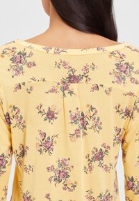 Gelbes Blusenshirt mit weitem Rundhalsausschnitt, leichtem Stoff, lila und grünen Blütenmustern sowie einem gerafften Falten-Detail auf der Rückseite.