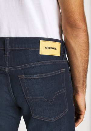 Man draagt donkerblauwe Diesel-spijkerbroek en toont achterzak en geel merkembleem, gekleed in wit overhemd met zichtbare arm.