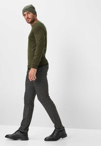 Camicia a maniche lunghe verde oliva abbinata a pantaloni grigi strutturati e scarpe stringate nere. L'uomo indossa un berretto verde a coste.