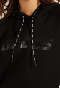 Sweat à capuche noir avec un logo "GUESS" brillant et des cordons avec embouts de marque. Le tissu a une texture lisse et unie.