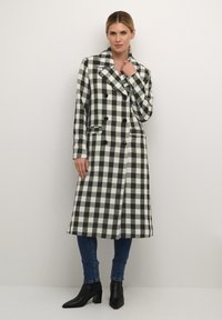 Abrigo largo a cuadros en blanco y negro con diseño de doble botonadura, grandes solapas y seis botones. Complementado con jeans oscuros ajustados y botas por el tobillo.