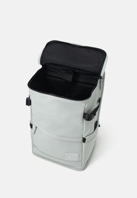 Mochila rectangular de color gris claro con parte superior abierta, interior negro, bolsillo frontal, correas laterales y etiqueta "Reutiliza Recicla Repite" en la parte delantera.