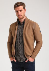 Cárdigan marrón con cremallera y tejido texturizado, que cuenta con un cuello alto y detalles en cremallera negra. Combinado con una camisa de botones con estampado debajo.