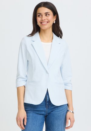 Vrouw glimlacht, draagt een lichtblauw blazer met opgerolde mouwen, wit topje, blauwe spijkerbroek en een zilveren armband tegen een effen achtergrond.