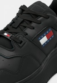 Black leather sneaker featuring textured accents, flat laces, and a branded patch with blue, red, and white details on the side.