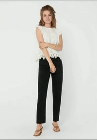 Weißes Spitzentop mit gewelltem Saum, kombiniert mit schwarzen geraden Hosen und braunen Sandalen. Das Modell steht mit übergeschlagenen Armen.