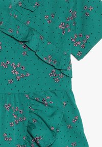 Zelenomodrá látková šaty s volánovými detaily, zdobené malými růžovými květy a černými puntíky. Měkká textura a vrstvený design.