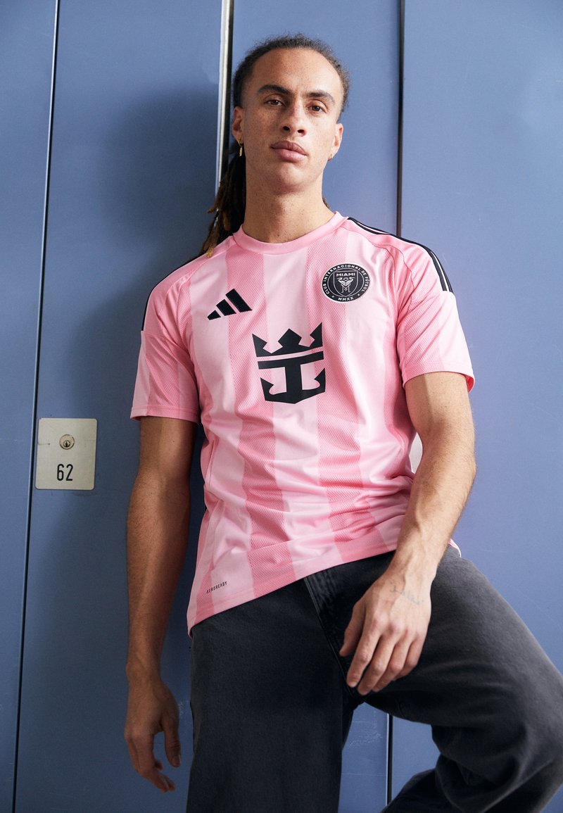 Rosa sportliches Trikot mit vertikalen Streifen, schwarzes Emblem mit einer Krone und einem Anker, adidas-Logo auf den Schultern, kombiniert mit dunkler Jeans.