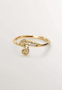 Anillo en tono dorado con diseño de caballito de mar, que presenta detalles texturizados y pequeñas piedras claras a lo largo de la banda, creando un acento caprichoso.