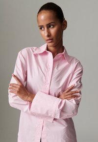 Šviesiai rožinė ilgomis rankovėmis medvilninė marškiniai su mygtukais, pasižyminti klasikiniu apykakliu ir laisvu kirpimu, su susuktomis rankogaliais.