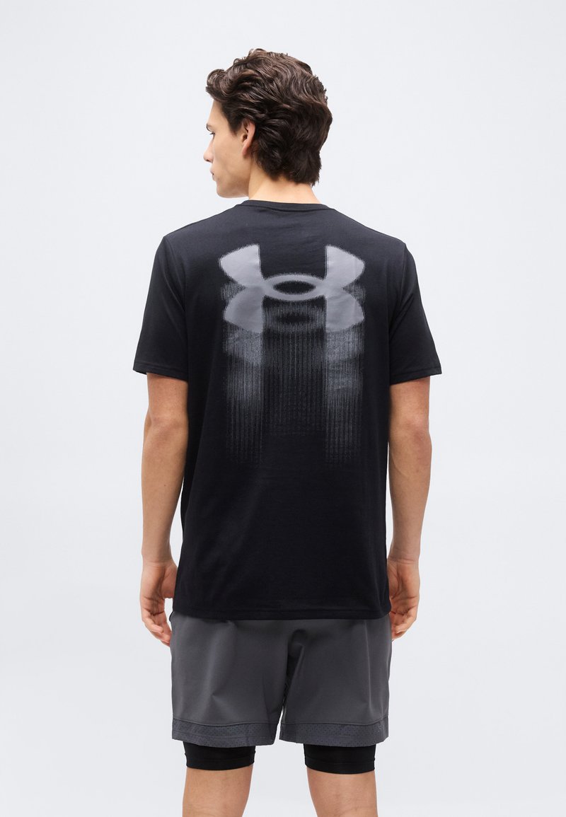 Mann trägt ein schwarzes Under Armour T-Shirt mit großem Logo auf der Rückseite und graue Shorts, steht mit dem Rücken zur Kamera vor einem schlichten weißen Hintergrund.