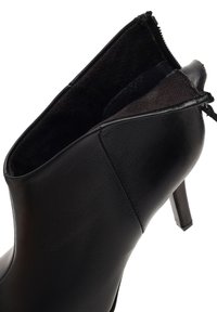 Botín negro hasta el tobillo con un exterior de piel suave, punta afilada, tacón alto delgado y cierre de cremallera, con una textura interior suave.
