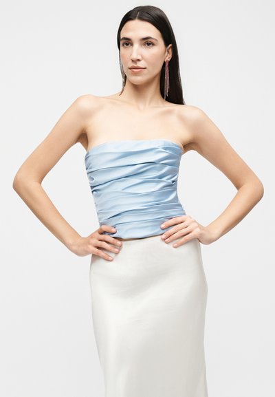 Γυναίκα με μακριά σκούρα μαλλιά που φοράει ένα αμάνικο φωτεινό γαλάζιο τοπ με ριχτά και λευκή φούστα, ποζάροντας με τα χέρια στη μέση μπροστά σε ένα απλό φόντο.