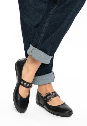 Schwarze Mary-Jane-Schuhe aus Leder mit silberner Schnalle und Ösen, getragen mit hochgekrempelten dunkelblauen Jeans ohne Socken.