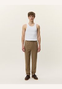 Camiseta sin mangas blanca, pantalones marrones con pliegues y zapatos oscuros. Los pantalones tienen una cinturilla elástica y una tela texturizada. Fondo neutro.