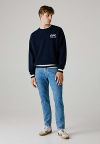 Granatowy sweter z białym logo, z prążkowanymi mankietami i dołem. W połączeniu z jasnoniebieskimi dżinsami i białymi trampkami z czarnymi akcentami.
