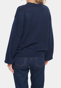 Pull oversize bleu marine avec poignets et ourlet côtelés, doté de détails en dentelle sur les épaules, associé à un jean en denim foncé.