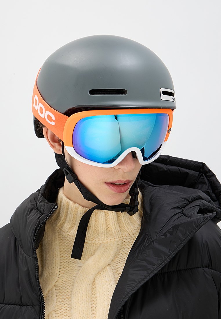 Szary kask z pomarańczowymi i białymi akcentami, w połączeniu z dużymi, lustrzanymi goglami narciarskimi. Osoba nosi czarną pikowaną kurtkę i kremowy sweter.