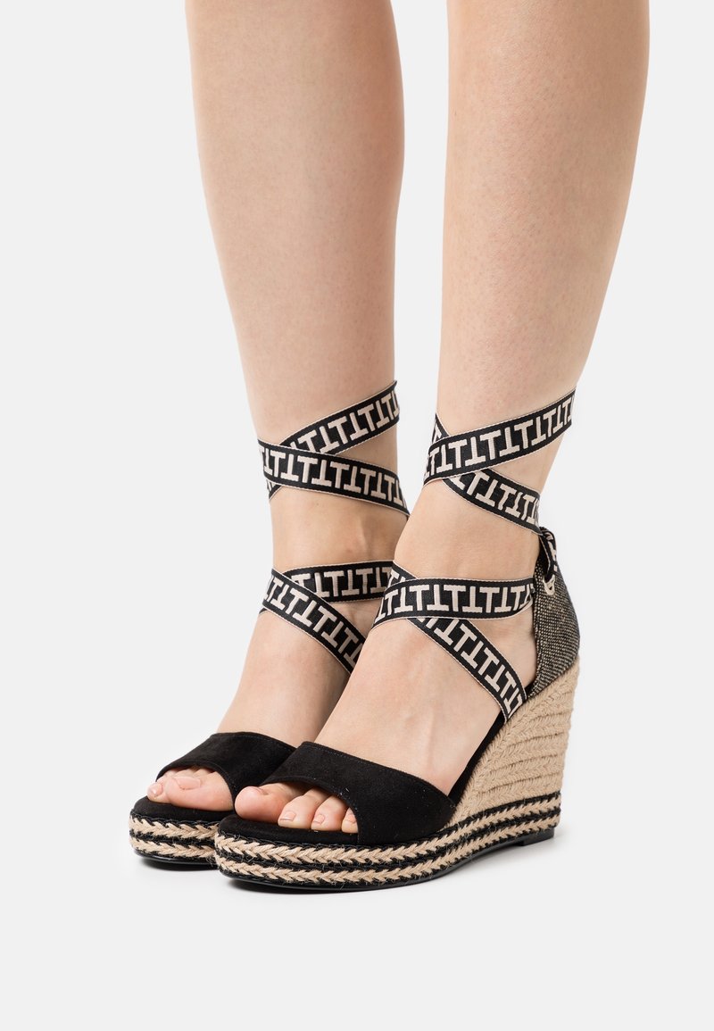Des pieds portant des espadrilles compensées noires avec des semelles en corde tressée et des straps autour de la cheville en noir et blanc présentant un motif en "T" répétitif.