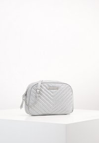 Borsa a tracolla argentata trapuntata con tracolla a catena, dotata di chiusura con zip, texture liscia e logo metallico decorativo sul davanti.