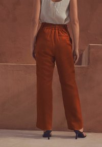 Pantalons amples de couleur rouille avec une taille élastique, associés à des sandales noires à talons. Texture lisse, design à jambes droites.