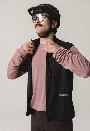 Mann mit schwarzem Fahrradhelm und Brille passt schwarze Weste über ein staubig rosa langärmeliges Shirt vor neutralem Hintergrund an.