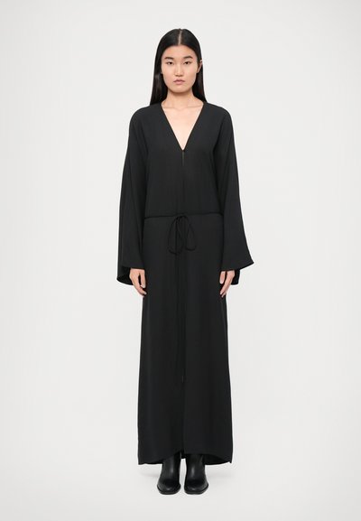 Schwarzes Maxikleid mit V-Ausschnitt und langen Glockenärmeln, mit einem Kordelzugbund und einer glatten, fließenden Stofftextur. Getragen mit schwarzen Stiefeln.