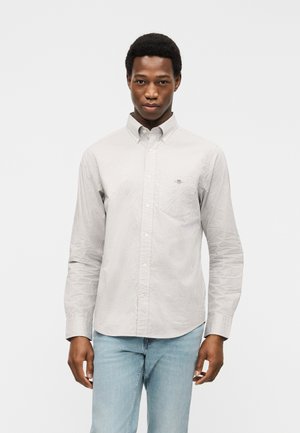 Jeune homme portant une chemise à carreaux grise claire à manches longues et un jean bleu clair, debout devant un fond blanc uni.