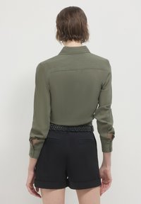 Camicia a maniche lunghe verde oliva con tessuto testurizzato, dettaglio con yoke posteriore e polsini con bottoni; abbinata a pantaloni corti neri a vita alta e una cintura intrecciata.